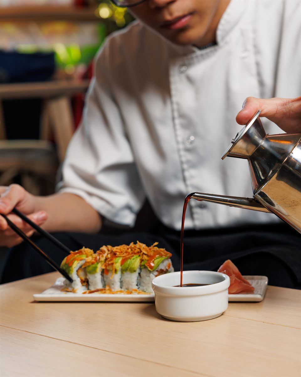 A chef pouring sauce beside a freshly plated sushi roll