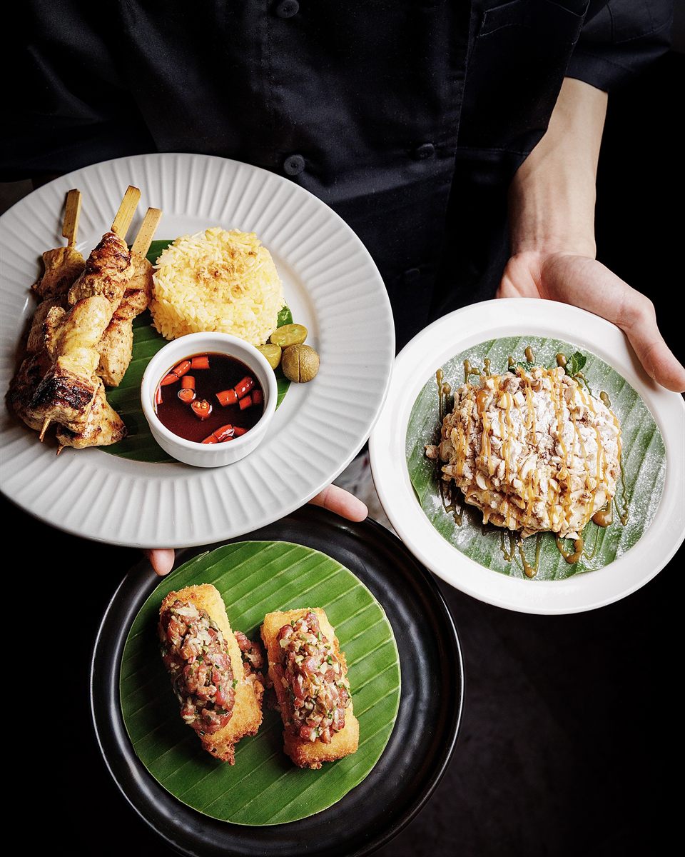 Overhead trio of grilled skewers, rice and banana-leaf plates held by a chef